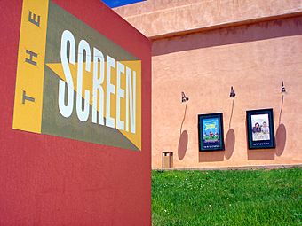 A sign in the foreground reads 'the Screen' while in the background an exterior view of a large, boxy building styled in pseudo-adobe stucco shows off a grassy lawn, a trash can, and lamp-illuminated movie posters