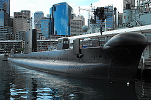 A submarine tied up alongside a wharf on a calm day. Numerous skyscrapers are in the background