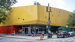 The facade of the Brooklyn Children's Museum building, which consists of a glass wall at street level and a yellow roof above it. A traffic light is in front of the entrance.