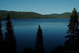 A large lake under blue skies surrounded by forested hills with steep slopes under a clear blue sky