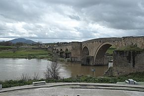 Photo of an old arched stone bridge over a river. There is a mountain in the distance