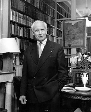 an older white man wearing a dark suit stands in an interior in front of bookshelves and a painting