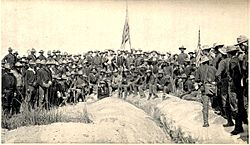 A black and white photo of US Army soldiers on 3 3 July 1898, in an upside down V type formation on top of Kettle Hill; two American flags are center and right. Soldiers facing camera.