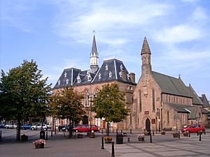 A square with three deciduous trees visible in the foreground. At the back of the square is the Town Hall, a neo-gothic style building. The town hall has two visible stories externally, together with a pitched roof with a number of dormers. In the centre of the roof area is a spire with a clock at its base. To the right of the Town hall is St Anne's church. The front of the church has a cylindrical cone spire.