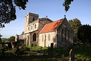A stone church seen from the southeast.  The chancel has a red tiled roof, the larger nave with clerestory has a battlemented parapet, and the tower has a plain parapet