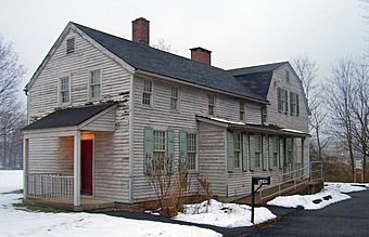 A two-story gray wooden house with light green shutters seen in winter, with snow on the ground. A long wing with two brick chimneys projects towards the left from a higher rear section. At lower right is a black mailbox with "5Ave" on it next to a clear paved driveway