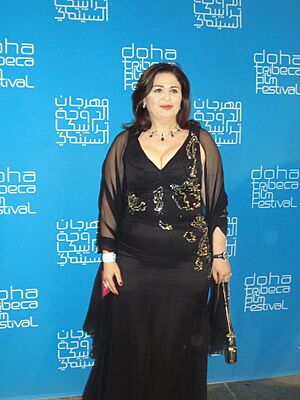 Elham Shahin wearing a black outfit, standing in front of Doha Tribeca Film Festival background, smiling and looking left of camera while holding an object, possibly a trophy, in left hand