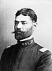 Head and shoulders of a man with neatly combed hair and a handlebar mustache wearing a plain military jacket with the letters "U.S.V." on the upright collar.
