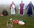 Four figures in medieval period costume stand outside on a grassy area. The image is misty.