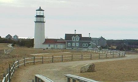 The lighthouse is in the background of the photo, and in the foreground is a large rock, or boulder. In the far distance, on the right, is the Pilgrim Monument tower in Provincetown.