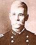 Head of a white man with a wide pointed mustache, wearing a dark double-breasted military jacket with the letters "U.S.V" on the collar.