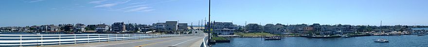 Panorama of town showing houses along the length of the image. A road descends from a bridge in the center to ground before terminating at a signalized intersection. An ocean is located in the background in the center of the image.