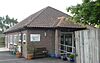 Three-quarter view of a small brick building with a brown tiled roof.  There are six windows in the side elevation; at the front is a central entrance porch formed by a projecting section of tiled roof supported on two thin brick columns.  A wooden fence partly hides the right-hand side of the building.