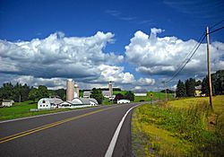 Country road with blue sky and clouds rolling overhead. There is a farm on the left and a sign indicating an intersection on the right.