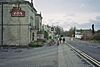 Gray stone building on the left with a pub sign outside it. A road is central to the picture with a white coloured building on the right.