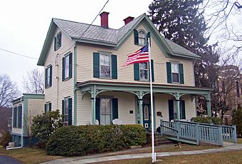 A pale yellow two-story wooden house with a pointed roof and ornate green trim. An American flag flies in front