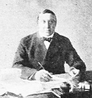 Black and white portrait of a man seated behind a table, with a pen in his right hand.