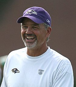 Color head-and-shoulders photograph of well-tanned, smiling white man with salt-and-pepper goatee (Chuck Pagano), wearing a white crewneck shirt and purple baseball cap, both emblazoned with Baltimore Ravens logos.