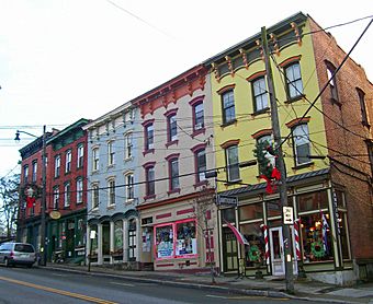A row of five brick buildings on a street ascending gently uphill to the left of the image, viewed from downhill to the right. All are three stories high with glass storefronts on the sidewalk and a flat roof. They are colored, from right, yellow, light brown, blue, red and unpainted red brick. There are Christmas wreaths on the two telephone poles in front.