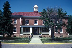 A large brick building dominates the photo. It has a large front porch with stairs leading to the main entrance, and a large spire atop the roof. Two trees in the front yard obscure part of the building.