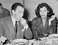 A man and woman sit next to each other at a table. The man eats while the woman smokes a cigarette.