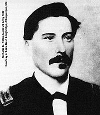 Black and white picture of a white male with a full dark mustache and squared goatee facing the camera and looking slightly to the right: He has dark, swarthy hair neatly combed over a broad forehead. His dress uniform has a white collar with the front ends turned down with a small bow tie.