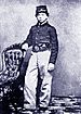 A white adolescent boy standing with his right arm resting on the back of a chair. He is wearing a forage cap, a dark military jacket, and light-colored pants.
