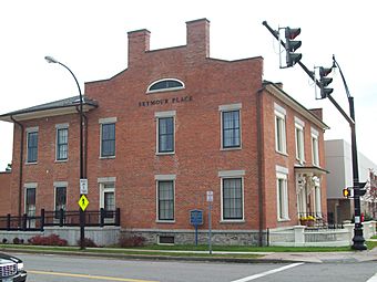 A brick building on a corner with a traffic signal in front of it. Two chimneys rise from a section that rises above the roofline on the side facing the camera; the front faces the right side. Metal lettering on the section facing the camera spells out "Seymour Place".