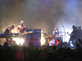 Four men are performing in front of an audience. The man at left is shown in right profile and is crouched over a keyboard with a second keyboard to his right. The second man is playing a guitar and singing into a microphone. The third man is seated behind a drum kit. The fourth man is shown in left profile playing a bass guitar. Two large speakers are behind the second man. Lights shine from the rear stage area.