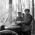 During additional refresher training, a Finnish soldier is having his breakfast served into a mess kit by another soldier from a steaming field kitchen in the forests of the Karelian Isthmus. More soldiers, two of them visible, are waiting in line for their turn behind him. It is early October and the snow has not set in yet.