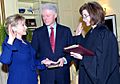 Hillary Clinton taking oath as Secretary of State on January 21, 2009. She is on the left side of the image, facing toward the right. The oath is being administered by Associate Judge Kathryn Oberly, who is standing directly in front of Hillary (on the right side of the photo) and facing toward the left. Bill Clinton, who is standing on both women's side in the background of the image, is holding a Bible.