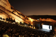 A shot of an outdoor amphitheatre taken at dusk, looking down toward a brightly lit stage. Large red cliffs are visible in the background, sloping down to the right. Several hundred people are visible between the camera and the stage.