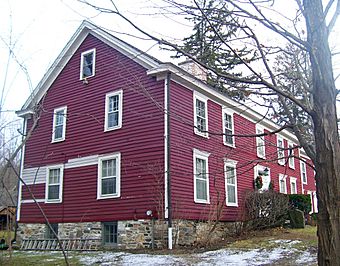 A red wooden house with some white siding seen from the side. There are bare trees and some snow in front.