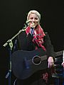Dianna Agron, smiling in a warm coat and scarf, holds a microphone and a black guitar while on a stage.