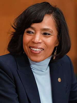 Alsobrooks smiling and looking to her left on a brown background, wearing a light blue shirt and black jacket with a U.S. Senate lapel pin.