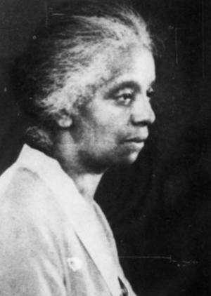 An older Black woman, grey hair dressed to the nape, wearing a light-colored jacket or dress
