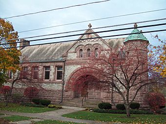 A stone building in two different shades of light brown behind some telephone wires and trees showing autumn color. On its right corner is a pointed roof in light green. Not too far towards the center is an entrance section with a pointed roof and wide round arched entrance