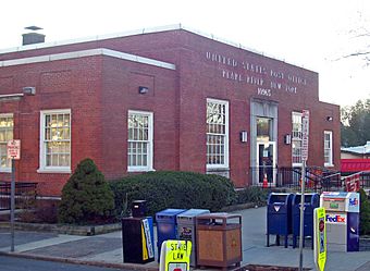 A brick building with a large main block and two smaller wings, seen from its left. Across the front metal letters spell out "United States Post Office, Pearl River, New York 10965". In the foreground are assorted mailboxes, private carrier drop boxes and newspaper boxes.