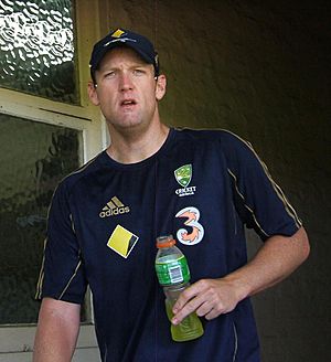 A man is peering in front of him, holding a drinks bottle with a yellow liquid in. He is wearing a dark blue t-shirt with gold piping, and four logos on it. He is also wearing a baseball cap of the same colours.
