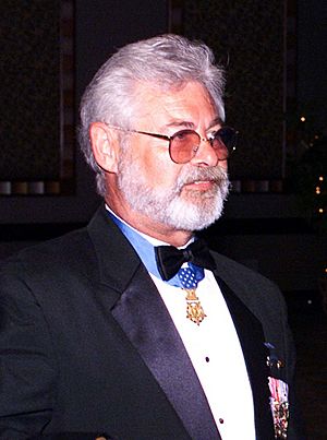 Head and shoulders of a man with gray hair, a full gray beard, and tinted glasses in a tuxedo. A medal, hanging from a blue ribbon around his neck, sits just below his bow tie.