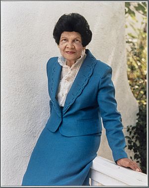 A woman with dark hair wearing a blue suit, leaning on a railing.