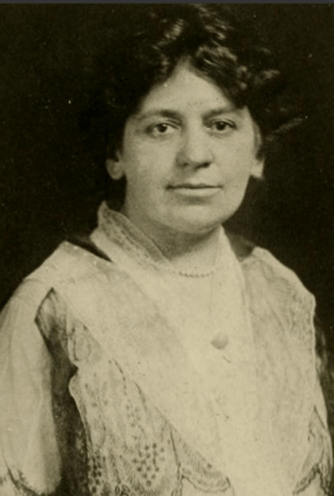 A white woman with dark curly hair, wearing a lace-trimmed dress or blouse