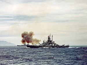 Color photo of a warship at sea. Smoke is rising from the bow of the ship, and land is visible in the background.
