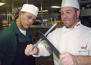 US Navy 031204-N-1711I-001 Mess Management Specialist 3rd Class Benjamin AlmodovarPacheco from Ceiba, Puerto Rico, watches carefully as culinary arts instructor Chef Sam Glass trains him on the best way to sharpen a butcher's