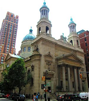 A stone building with a pediment and columns on the main entrance, two towers with green rounded tops and columns, and a dome at the rear lit by late afternoon sun from the right. There are traffic lights in front.