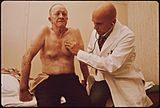 A bald physician listens with a stethoscope to the chest of an old, bare-chested man