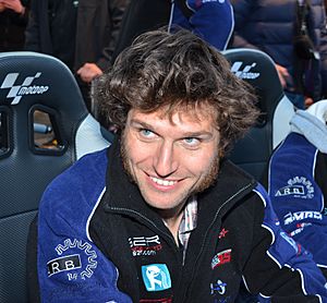 Man with wavy dark hair wearing a blue top smiling and looking at something off camera