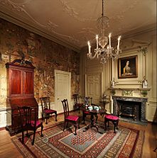 Photo of a richly decorated drawing room with a table in the middle set for tea. A chandelier is lit and there is a painted portrait of a woman on the chimney-breast