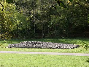 Elevated view of the cairn in the middle distance, from its side, with deciduous trees in leaf to its rear. To its front passes a wide asphalt path, dissecting flat ground of short grass. The tumulus' trapezium shape is evident, its boulders retained by a short wall, missing at the very front, left, where the rubble has tumbled out.