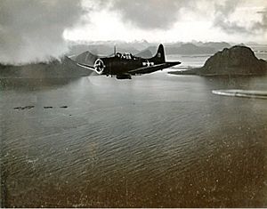 Black and white photo of a single-engined monoplane aircraft flying over a body of water. Steep mountains rise out of the water in the background.